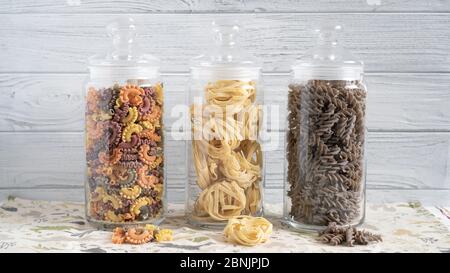 Different types of pasta in jars on white wooden backgraund. Colored creste di gallo, fettuccine and dark torti. Stock Photo
