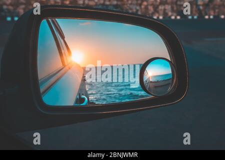 Reflection of the beautiful sunset above the sea through the car's wing mirror in the parking lot close to the sea Stock Photo