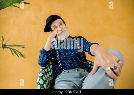 Handsome cheerful trendy senior man with well-groomed beard, wearing dark blue shirt, suspenders, gray pants, and black hipster cap, sitting in chair Stock Photo