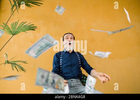 Studio portrait of excited joyful handsome elderly man in stylish clothes, with well-groomed beard, and open mouth, which is sitting under paper money Stock Photo