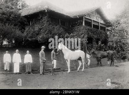 British man with his servants and horses outside his bungalow, late 19th century SE Asia, possibly Singapore Stock Photo