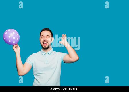 A handsome man holds a balloon in his hand and rejoices and screams at a holiday or birthday. A man inhaled laughing gas, helium and laughs on a blue Stock Photo