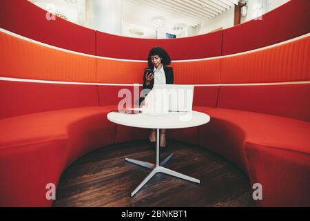 Wide-angle shot of a young charming African-American businesswoman sitting on a curved sofa of red color in an office lounge and using her phone; lapt Stock Photo