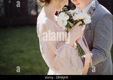 The bride and groom hug, stand facing each other. The bride in a pink dress holds in her hands a bouquet of white and pink peonies. The bouquet is tie Stock Photo