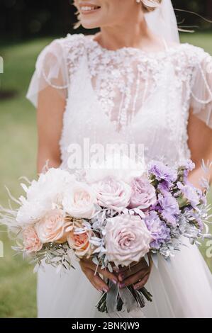 Close-up of a bride’s bouquet of roses, carnations in white, pink, violet, lavender, peach shades. The bride in a lace white dress holds a bouquet