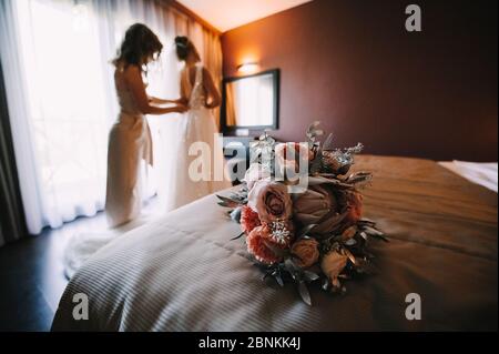 in the foreground a bouquet of roses, carnations, protea in white, pink, violet, lavender shades; against the background, mom buttons the wedding dres Stock Photo