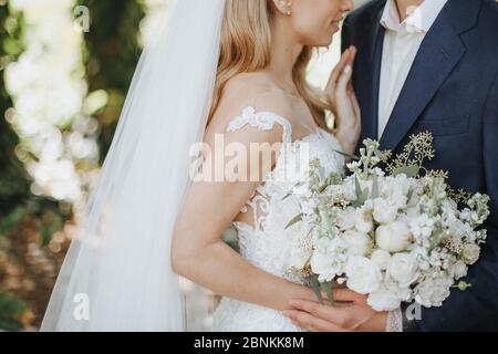 Close-up of a bride’s bouquet of white flowers. The bride in a lace white dress holds a bouquet in her hands. The bride and groom hug, stand facing ea Stock Photo