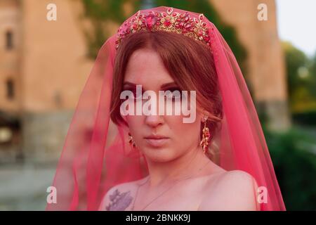 Portrait of attractive redhead tattooed woman in long red dress, diadema and red veil on blurred medieval castle background Stock Photo