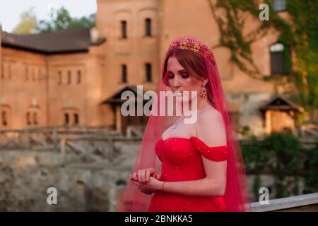 Portrait of attractive redhead tattooed woman in long red dress, diadema and red veil on blurred medieval castle background Stock Photo