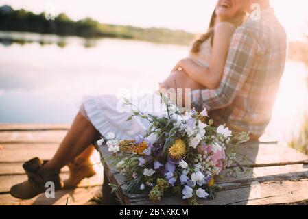 in the foreground a bouquet of white and lilac bells; against the background, a guy and a pregnant girl are sitting on the pier embracing Stock Photo