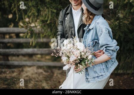 The bride in a denim jacket and hat holds a bouquet in white and purple shades. Groom hugs her from behind Stock Photo