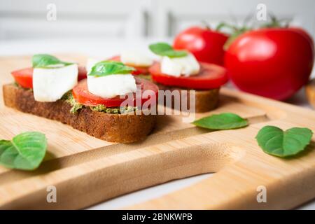 Traditional Italian caprese bruschetta: sandwich toast with basil pesto sauce, mozzarella cheese and tomatoes on the wooden board surrounded by basil l Stock Photo