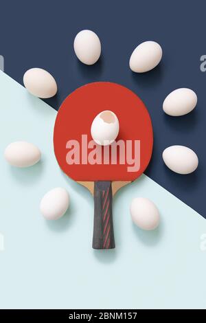 The red ping pong racket is in the center. There is a broken egg on the racket. There are whole eggs lying around. The background color is blue. Stock Photo