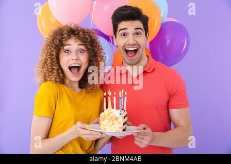 Image of excited couple man and woman celebrating birthday with multicolored air balloons and piece of cake isolated over violet background Stock Photo