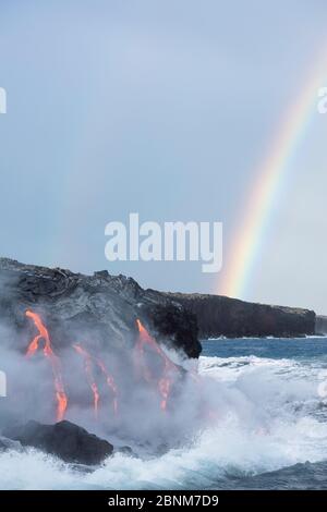 Hot lava from the 61G flow, emanating from Pu'u O'o on Kilauea Volcano, flows through lava tubes into the ocean in front of a double rainbow at the Ka Stock Photo