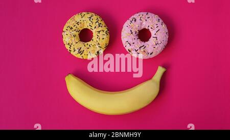 Two donuts and a banana lie on the pink surface, forming an abstraction of the emoticon - smile. Taken from the top point Stock Photo