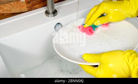 Female hands wearing yellow rubber gloves washing white plate in kitchen sink with foam sponge. Stock Photo