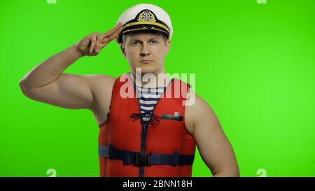 Young muscular sailor man works as lifeguard at beach salutes with hand to camera. Seaman guy life saver in sailor's vest, lifejacket, captain nautical hat. Isolated on chroma key background Stock Photo