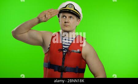 Young muscular sailor man works as lifeguard at beach salutes with hand, looking up. Seaman guy life saver in sailor's vest, lifejacket, captain nautical hat. Isolated on chroma key background Stock Photo