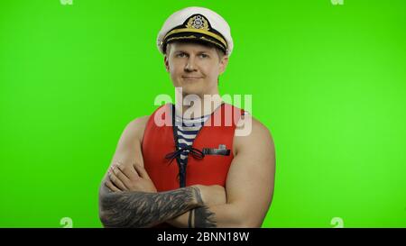 Young muscular sailor man works as lifeguard at beach arms crossed, looking at camera. Seaman guy life saver in sailor's vest, lifejacket, captain nautical hat. Isolated on chroma key background Stock Photo