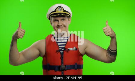 Young muscular sailor man works as lifeguard at beach show thumbs up and looking at camera. Seaman guy life saver in sailor's vest, lifejacket, captain nautical hat. Isolated on chroma key background Stock Photo