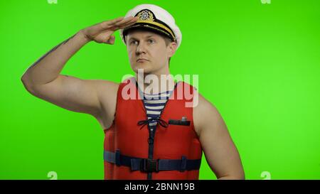 Young muscular sailor man works as lifeguard at beach observe the territory. Seaman guy life saver in sailor's vest, lifejacket, captain nautical hat. Isolated on chroma key background. Green screen Stock Photo