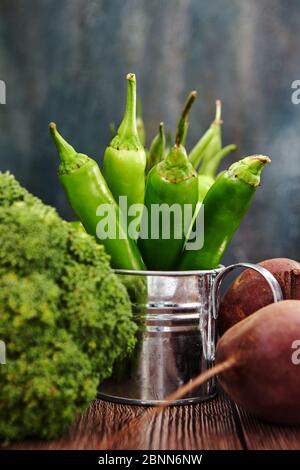 Green hot pepper in an enameled watering can, broccoli and beets on wooden background. Stock Photo