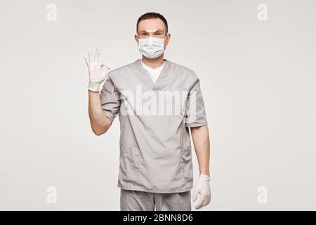 Young man in medical uniform, protective mask and glasses showing OK gesture with one hand and posing at the white background, isolated. Healthcare Stock Photo