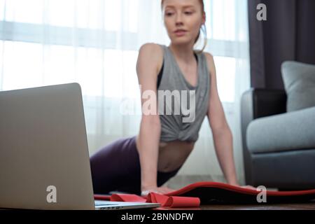 Slender girl in sports uniform is engaged in stretching at home, looking at the laptop, focus on the laptop Stock Photo