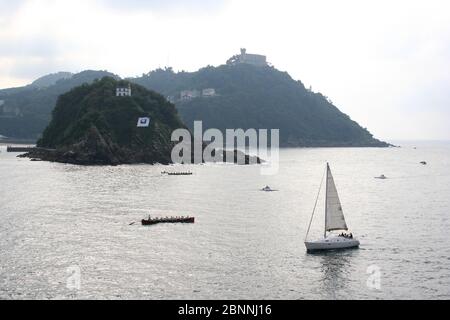 Boat race festival with view towards island racing row boats and yacht in full sail Basque week for the Flag of La Concha San Sebastian Gipuzkoa Spain Stock Photo