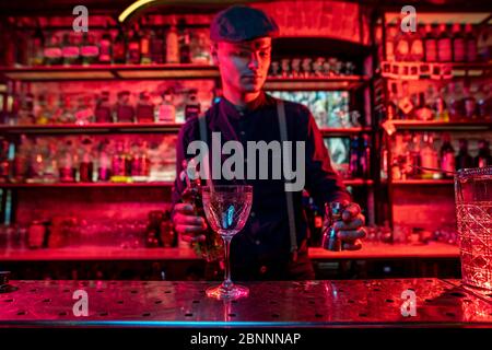 Professional barman preparing the alcoholic cocktail with shot in multicolored, deep red neon light, gives it to guest. Entertainment, drinks, service concept. Modern bar, trendy neoned colors. Stock Photo