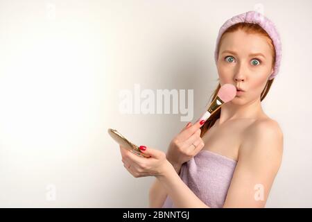 A redhead girl is standing on a white background in a towel, holding a small mirror and opening eyes wide, hiding behind a brush Stock Photo