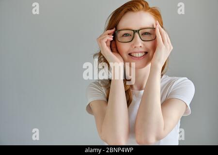 A red-haired girl in a white T-shirt stands on a gray background and smiles as she holding onto her glasses. Stock Photo