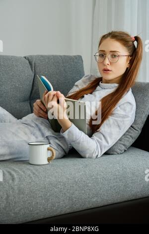 A girl in glasses and pajamas is lying on the couch with a book Stock Photo
