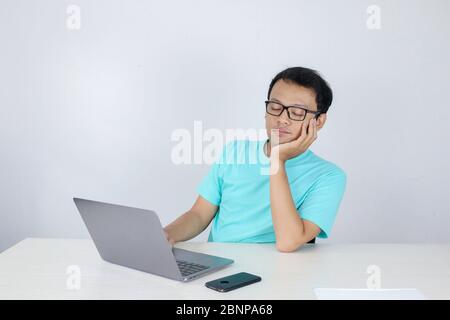 Young Asian Man is Lazy and tired with sleep when working in the front of Laptop. Indonesian man wearing blue shirt. Stock Photo