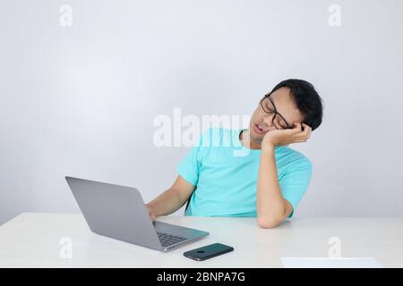 Young Asian Man is Lazy and tired with sleep when working in the front of Laptop. Indonesian man wearing blue shirt. Stock Photo