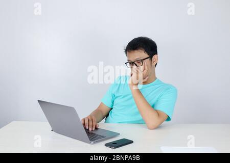 Young Asian Man is Lazy and tired with sleep when working in the front of Laptop. Indonesian man wearing blue shirt. Stock Photo