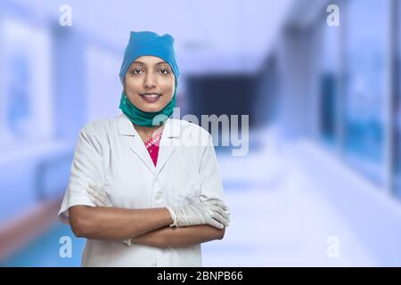 Happy Young Female Medical Worker,Nurse Standing Cross Arms With Hospital Corridor In The Background. Covid-19, Coronavirus Pandemic. Stock Photo