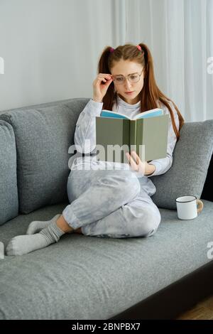 Red-haired girl in pajamas sits on the couch with her legs tucked and reads a book, adjusting her glasses Stock Photo