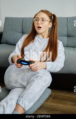 Redhead girl in glasses and pajamas sits on the floor with a game joystick, enthusiastically opening her mouth Stock Photo