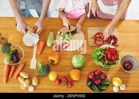 Family cooks fresh vegetables on the table in the kitchen. Hands of mother father and daughter with a knife are preparing a vitamin salad. No faces. T Stock Photo