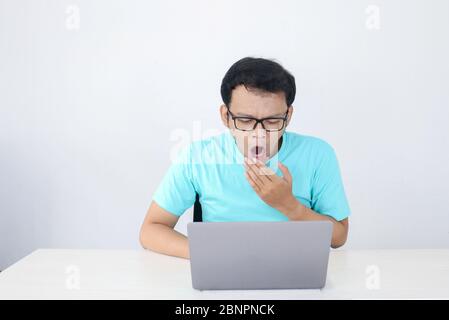 Young Asian Man is Lazy and tired with sleep when working in the front of Laptop. Indonesian man wearing blue shirt. Stock Photo