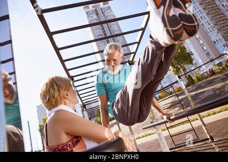 Smiling mature man in a great shape doing push ups on parallel bars and talking with his wife. Healthy mature couple doing sport together in the Stock Photo