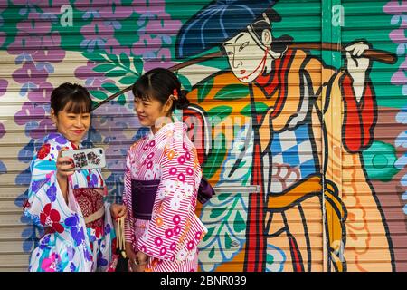 Japan, Honshu, Tokyo, Asakusa, Two Women in Kimono Taking Selfie Photos in front of Colourful Store Shutter Painting Stock Photo
