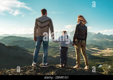 Family of father, mother and young child stands in mountains and looks at sunset Stock Photo