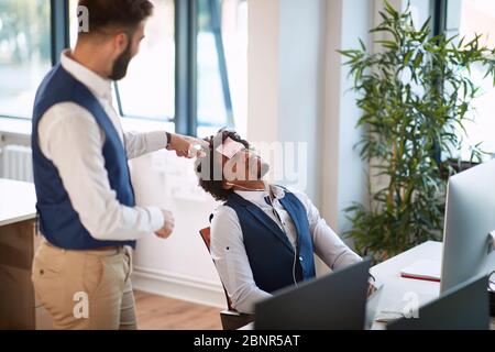 Coworkers having fun in the office. Putting a sticky note on a coworker prank. Friendly businessmen being silly. Stock Photo