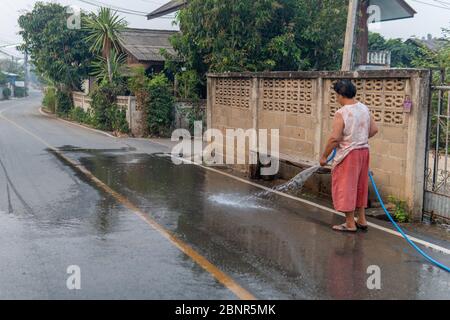 Thai lady hosing off her portion of the street to prevent dust in the city of Pai Stock Photo