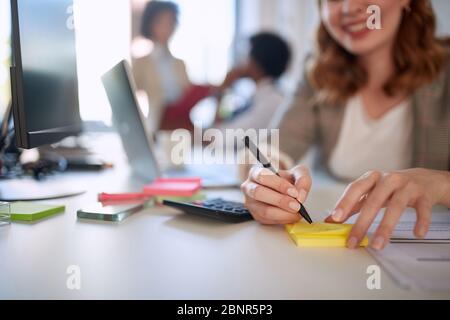 Businesswoman writing on a stickynote. Smiling woman taking notes at work. At the desk closeup. Stock Photo