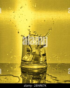 Ice cubes splashing into glass of water. Yellow background Stock Photo