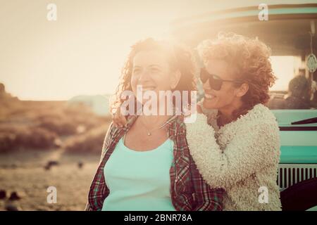 Couple of cheerful happy women adult friends enjoy together the outdoor leisure activity having fun and smiling - blue vintage van in background for travel concept - sunset backlight Stock Photo